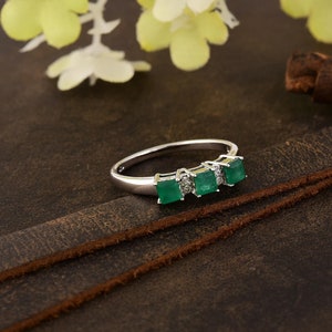 May include: A silver ring with three emerald green gemstones set in a row. The ring is on a brown leather surface.