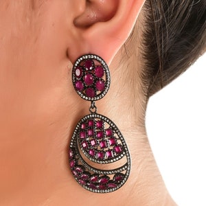 May include: A pair of dangle earrings with a black metal setting and pink gemstones. The earrings feature a round top and a teardrop-shaped bottom.