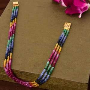 May include: A multi-strand necklace made of colorful gemstones. The necklace is made of four strands of beads, each strand featuring a different color of gemstone. The gemstones are red, green, blue, and yellow. The necklace is finished with gold clasps.