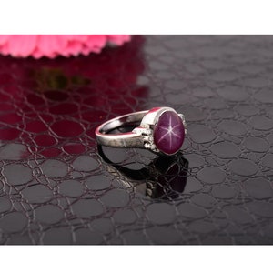 Op de afbeelding: Een zilveren ring met een grote, ovale, rode edelsteen met een stervormig patroon. De ring ligt op een zwart, getextureerd oppervlak.