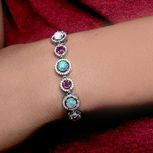 Puede incluir: Un brazalete de plata con una fila de piedras preciosas redondas y coloridas engastadas en un bisel de plata. Las piedras preciosas están rodeadas de piedras preciosas más pequeñas y transparentes. El brazalete está en la muñeca de una persona.