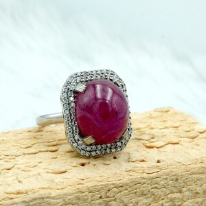 May include: A silver ring with a large, cabochon-cut ruby surrounded by a halo of smaller diamonds. The ring is set on a wooden surface.