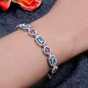 Może przedstawiać: Srebrna bransoletka z naprzemiennymi niebieskimi i różowymi kamieniami szlachetnymi. Bransoletka jest ozdobiona małymi przezroczystymi kamieniami wokół każdego kamienia szlachetnego.