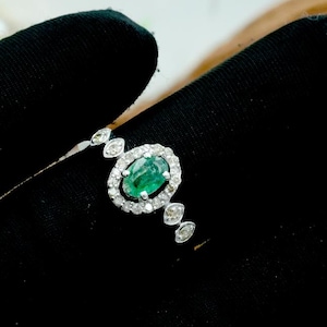 May include: A silver ring with an oval emerald gemstone, encircled by small diamonds. Further diamond accents are set along the band. The ring is shown against a black backdrop.