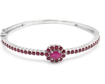 May include: A silver bangle bracelet featuring a central cluster of red gemstones, encircled by smaller red gems. The bracelet has a hinged clasp. The gemstones are a deep red colour, set against a white backdrop.