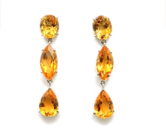 May include: A pair of dangling earrings with three yellow-orange gemstones each. The top stone is oval, the middle is marquise-shaped, and the bottom is a teardrop. The earrings are set in silver metal and hang against a white background.
