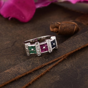 May include: A silver ring with a row of four square-shaped gemstones. The gemstones are emerald green, ruby red, diamond white, and sapphire blue.