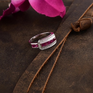 May include: A silver ring with a rectangular setting featuring a row of red ruby stones and a border of smaller white diamonds.