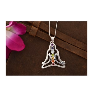 Puede incluir: Collar con colgante de plata que presenta una figura estilizada en una postura de meditación sentada. La figura está adornada con siete piedras preciosas de colores que representan los siete chakras.