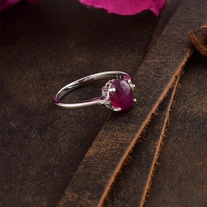 Peut inclure: Une bague en argent avec une grande pierre de rubis ovale entourée de petites pierres claires.