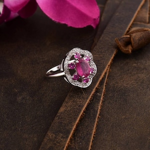 May include: A silver ring with a floral design. It has a large oval ruby in the centre, encircled by smaller pink gems and clear stones. The ring is set on a brown leather surface.