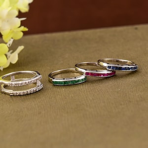 Könnte beinhalten: Vier Silberringe mit verschiedenen Edelsteinen. Die Ringe liegen auf einer braunen Oberfläche. Der Ring links hat eine doppelte Reihe von Diamanten. Die anderen Ringe haben eine einzelne Reihe von Edelsteinen. Die Edelsteine sind smaragdgrün, rubinrot und saphiblau.