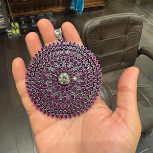May include: A large circular pendant featuring a central clear round stone surrounded by multiple concentric rings of small, teardrop-shaped magenta stones set in a dark metal frame with a silver-toned bail at the top for attachment to a chain.