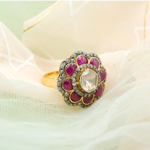 Op de afbeelding: Een gouden ring met een grote, heldere edelsteen omringd door kleinere roze edelstenen. De ring is gezet in een bloemvormig ontwerp.