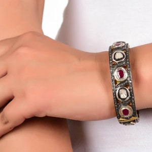 May include: A silver and gold bangle bracelet with red and white gemstones. The bracelet is set with small diamonds and has a delicate design.