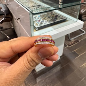 May include: A gold ring with three rows of gemstones. The top and bottom rows feature clear, small stones, while the middle row showcases rectangular, red gemstones. The ring is held in a person's hand, with a jewelry display case in the background.