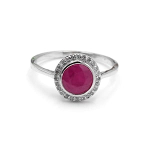 Op de afbeelding: Een zilveren ring met een grote, ronde, dieproze edelsteen, omringd door een halo van kleinere, heldere stenen. De ring heeft een dunne, gepolijste band, wat zorgt voor een klassiek en elegant ontwerp.