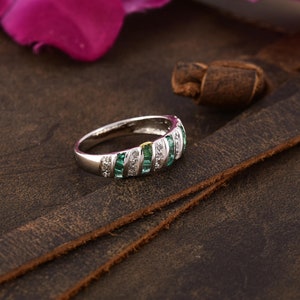 Puede incluir: Un anillo plateado con una serie de piedras verdes esmeralda y blancas. El anillo tiene una banda de piedras verdes esmeralda y blancas alternas. El anillo está sobre una superficie de cuero marrón.