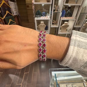 May include: A silver bracelet with oval-cut, deep red gemstones, each surrounded by smaller, clear stones. The bracelet is displayed on a wrist, showcasing its design and sparkle. The jewelry is set against a background of a jewelry store.