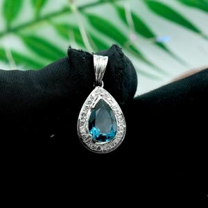 Op de afbeelding: Een druppelvormige hanger met een grote, diepblauwe edelsteen in het midden, omringd door een halo van kleinere, heldere stenen. De hanger is gezet in een zilverkleurig metaal en hangt aan een bijpassende balein. Zwarte en groene achtergrond.