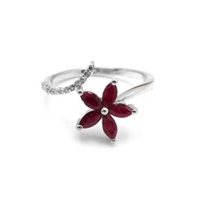 Może przedstawiać: Srebrny pierścionek z kwiatowym wzorem. Pierścionek ma kwiat z sześcioma głęboko czerwonymi płatkami. Zakrzywiony akcent jest inkrustowany małymi, przezroczystymi kamieniami. Pierścionek jest na białym tle.