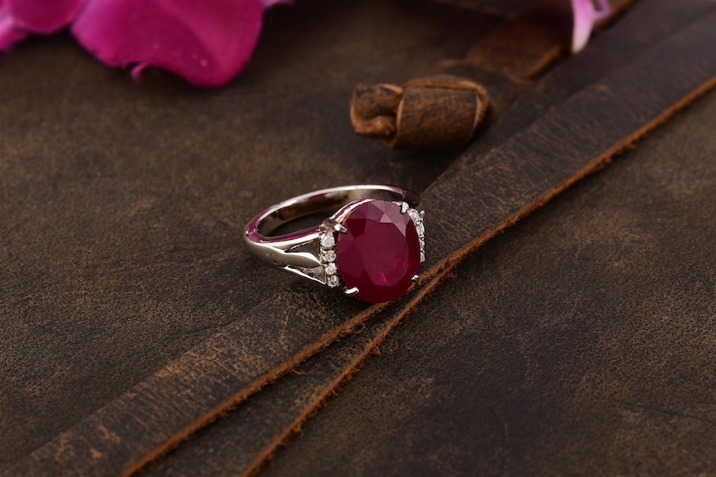 May include: A silver ring featuring a large oval ruby gemstone surrounded by smaller diamonds. The ring is displayed on a brown leather surface.