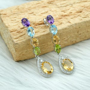 Puede incluir: Pendientes de plata con una cascada de piedras preciosas de colores. Cada pendiente tiene una amatista, seguida de piedras azules, amarillas y verdes, que culminan en una gran piedra amarilla en forma de lágrima. Las piedras están engastadas en un marco de plata.