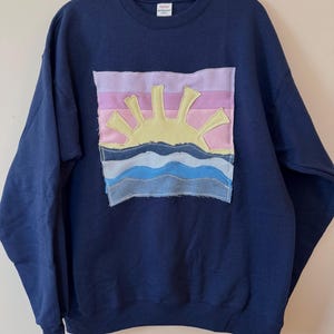 May include: Navy blue sweatshirt with a colorful, stitched-on patch featuring a yellow sun, pink and purple sky, and blue waves. The sweatshirt is hanging on a gray hanger.