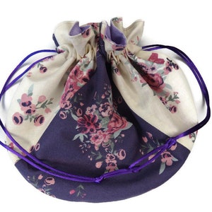 May include: A round drawstring pouch with a floral design. The pouch is split into cream and deep purple sections, each decorated with pink and purple flowers. Purple drawstrings close the top.