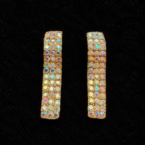 Puede incluir: Un par de pendientes rectangulares de tipo botón con tres filas verticales de pedrería iridiscente multicolor engastada en un marco de metal dorado. Las piedras muestran un brillo reflectante similar al arcoíris en sus superficies facetadas.