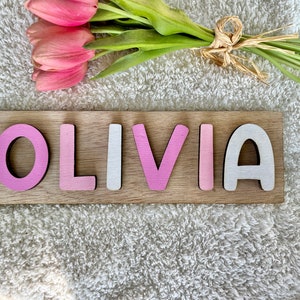 May include: Wooden name puzzle with the name "OLIVIA" spelled out in pink and white letters. The letters are on a light brown wooden base.