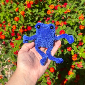 May include: A blue crocheted frog toy with black eyes. The frog is sitting in a hand, with a background of orange and yellow flowers.