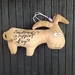 May include: A tan donkey-shaped plush toy with the words "Emotional Support Toy" written on its side. The toy has a rope tail, a white string for hanging, and is set against a dark wood-paneled background.