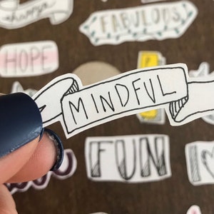 May include: A white sticker with a black banner that says "MINDFUL" in a handwritten font. The sticker is being held by a finger with blue nail polish.