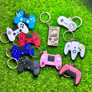 May include: A selection of vibrant video game controller keyrings in diverse colours, including blue, white, red, black, and pink. The keyrings are displayed on a green surface, highlighting their detailed designs and button layouts.
