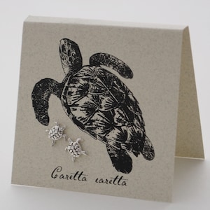 Peut inclure: Une paire de boucles d'oreilles en argent en forme de tortue sur une carte avec une illustration en noir et blanc d'une tortue de mer. La carte porte également le texte "Caretta caretta" imprimé dessus.