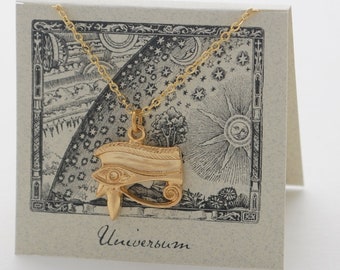 Könnte beinhalten: Goldfarbene Anhängerkette mit einem Horusauge-Design. Der Anhänger hängt an einer filigranen Kette und wird auf einer Karte mit einer Illustration von Himmelskörpern und dem Wort "Universum" präsentiert.