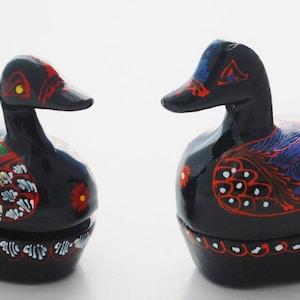 Può includere: Due statuine di anatra di legno dipinte di nero con motivi floreali colorati. Le anatre si guardano e hanno il becco aperto. Le anatre sono alte circa 10 cm.