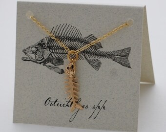 May include: A gold necklace featuring a fish bone pendant. The pendant is a detailed replica of a fish skeleton. The necklace is displayed on a card with a black and white illustration of a fish skeleton and the text "Osteichthyes".