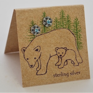 May include: Sterling silver snowflake earrings with light blue crystals on a brown card with a drawing of a mother bear and cub in a forest setting.