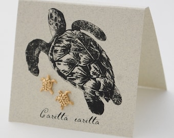 Op de afbeelding: Gouden schildpad oorbellen met een zwart-witte illustratie van een zeeschildpad op een kaart met de tekst "Caretta caretta".