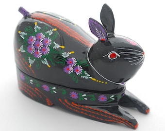 Könnte beinhalten: Schwarze Holzbox in Form eines Kaninchens mit einem Scharnierdeckel. Die Box ist mit bunten Blumendesigns in den Farben Lila, Grün, Gelb und Rot verziert.
