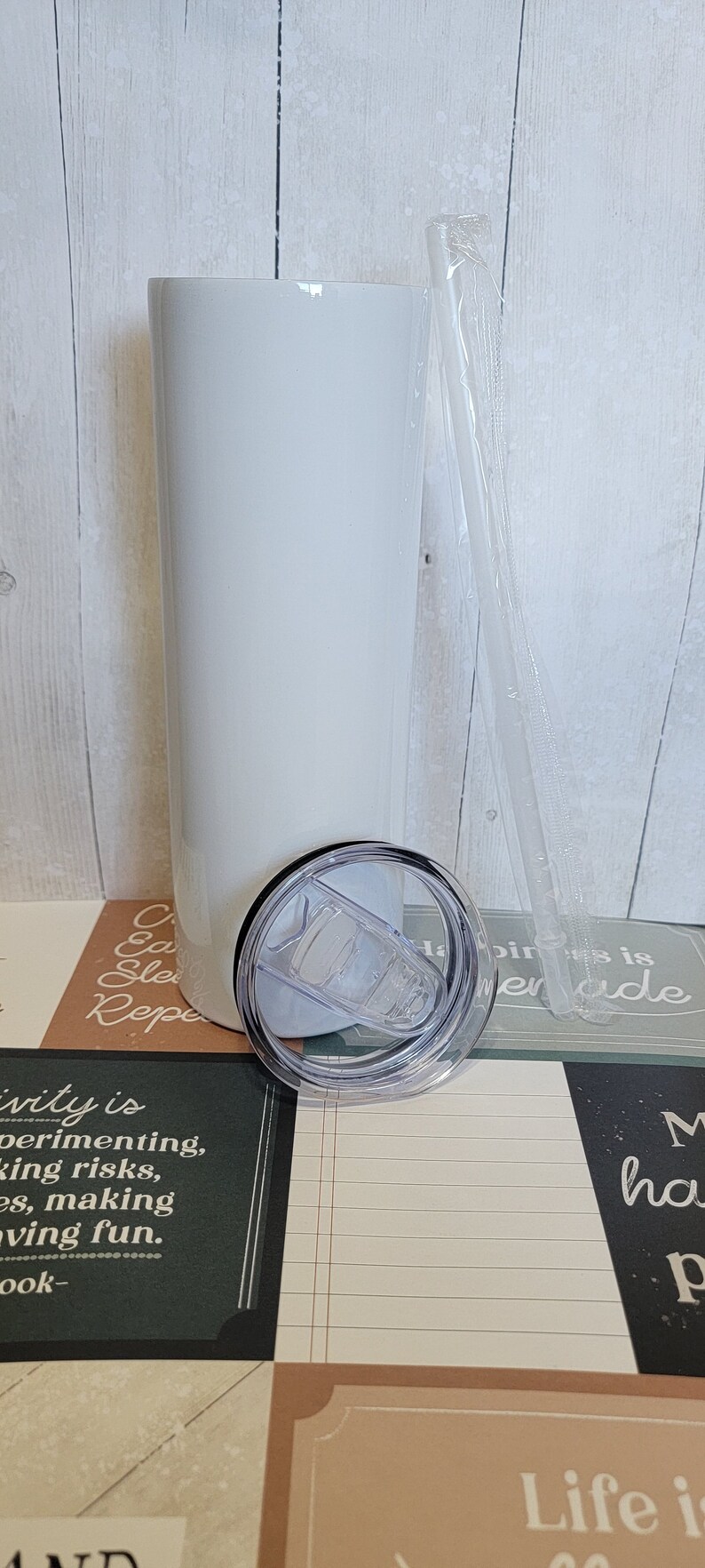 Op de afbeelding: Een witte roestvrijstalen tumbler met een doorzichtig deksel en een doorzichtige plastic rietje. De tumbler staat op een tafel met een groen en bruin gepatroneerd oppervlak. De tekst "Creativity is experimenting, taking risks, making, having fun." is zichtbaar op het oppervlak.