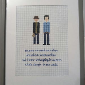 May include: Framed cross-stitch art featuring two figures in hats and dark pants. The figures are on a white background with blue text that reads "because we need each other...who's sleepin' in our souls."
