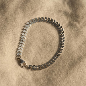 May include: A silver bracelet featuring a repeating chevron design. The bracelet is composed of small, flat, diamond-shaped links, forming a flexible chain. A small clasp secures the bracelet. The background is a tan fabric.