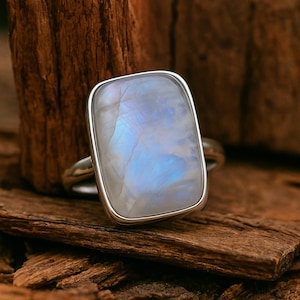 May include: A rectangular moonstone ring with a silver band. The moonstone has a pearly white base with blue and gray internal reflections. The ring is set on a piece of weathered wood.