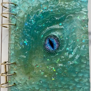 May include: A teal blue resin-bound journal with a gold ring binding. The cover features a large iridescent eye with a black pupil and a purple and pink iris.
