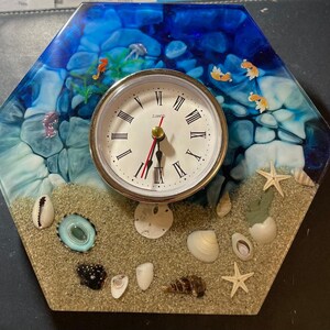 May include: A hexagonal resin clock with a blue and white ocean scene. The clock face is white with black Roman numerals and a gold-colored hour hand and minute hand. The clock is surrounded by seashells, starfish, and seahorses.