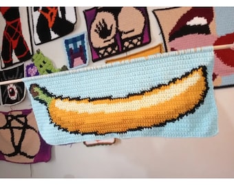 May include: A crocheted wall hanging featuring a yellow banana on a light blue background. The banana is made with yellow, white, and black yarn.