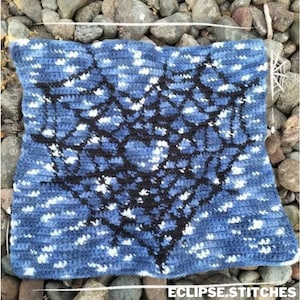 Op de afbeelding: Een blauw en wit gecrochet vierkant met een zwart geometrisch ontwerp in het midden. Het ontwerp lijkt op een spinnenweb.